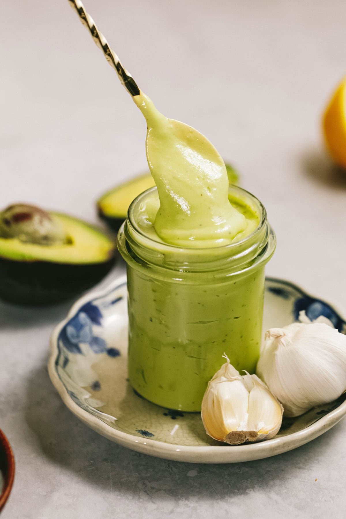 A small jar of creamy green sauce is displayed on a plate with a spoon dipped in it. Garlic cloves and half an avocado are placed nearby.