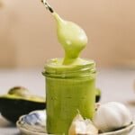 A hand holds a spoon dipping into a jar of green sauce, with garlic cloves and an avocado half placed beside the jar on a small plate.