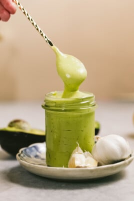 A hand holds a spoon dipping into a jar of green sauce, with garlic cloves and an avocado half placed beside the jar on a small plate.