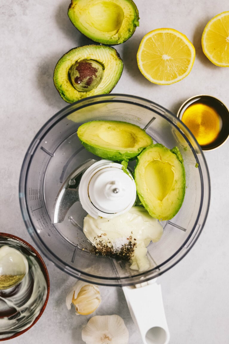 Ingredients including avocado, lemon halves, garlic, honey, yogurt, and spices are arranged around a food processor, which contains avocado, yogurt, and spices, on a gray countertop.