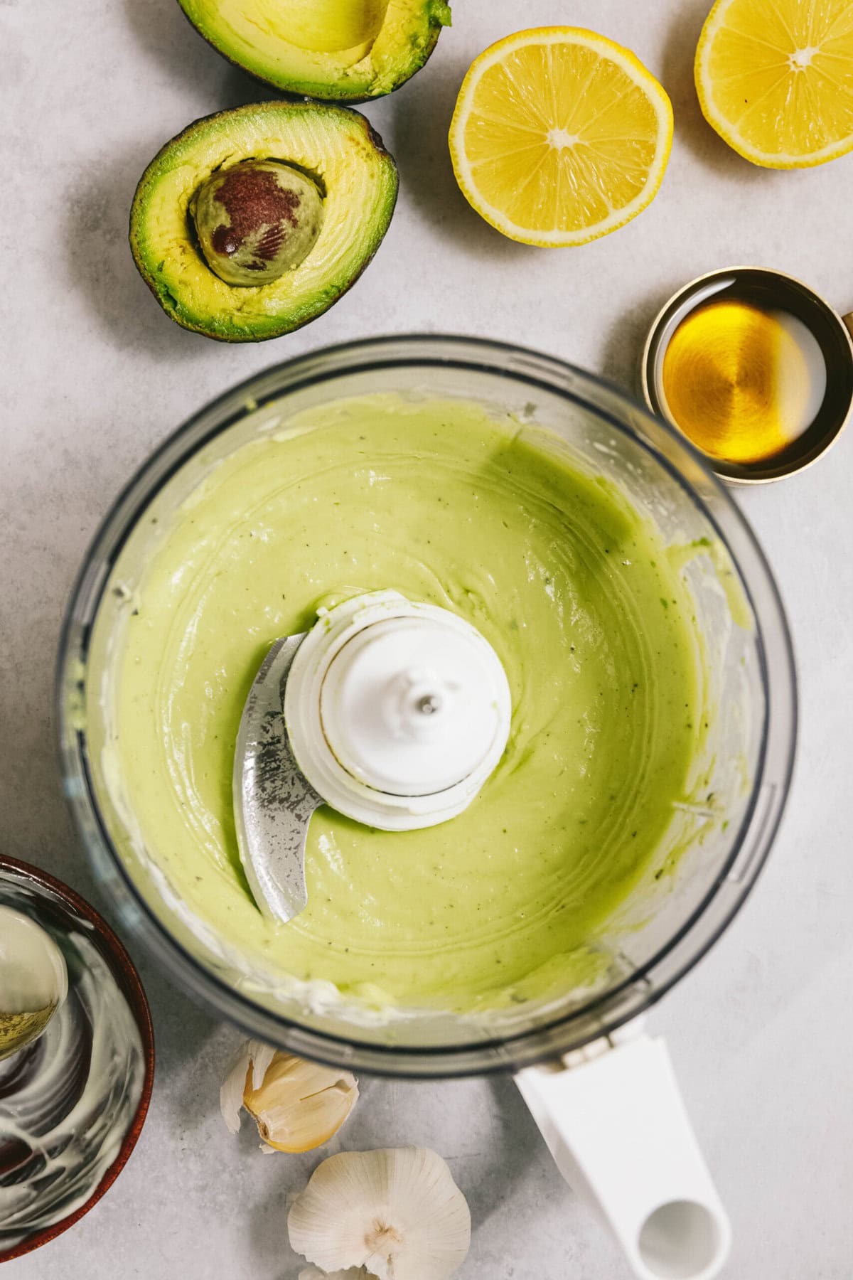 A food processor containing green avocado sauce is surrounded by avocado halves, lemon halves, garlic cloves, and a small dish of oil on a countertop.