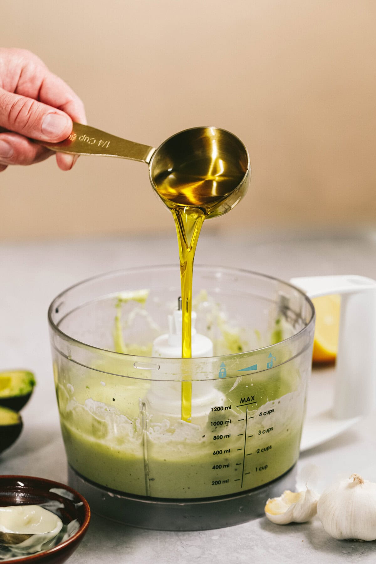 A hand pours olive oil from a measuring cup into a food processor containing a green mixture. An avocado, garlic, and lemon are nearby on the counter.