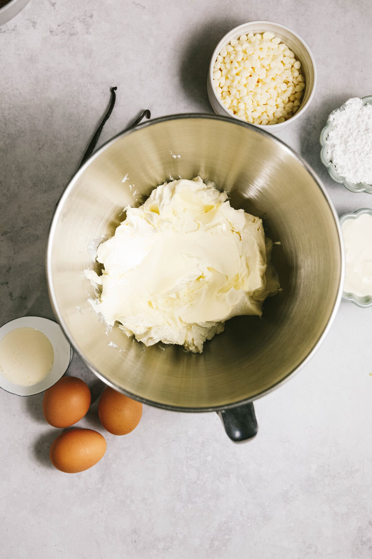 A metal mixing bowl containing whipped butter is surrounded by three eggs, a cup of heavy cream, a bowl of white chocolate chips, and a bowl of powdered sugar on a light gray surface.