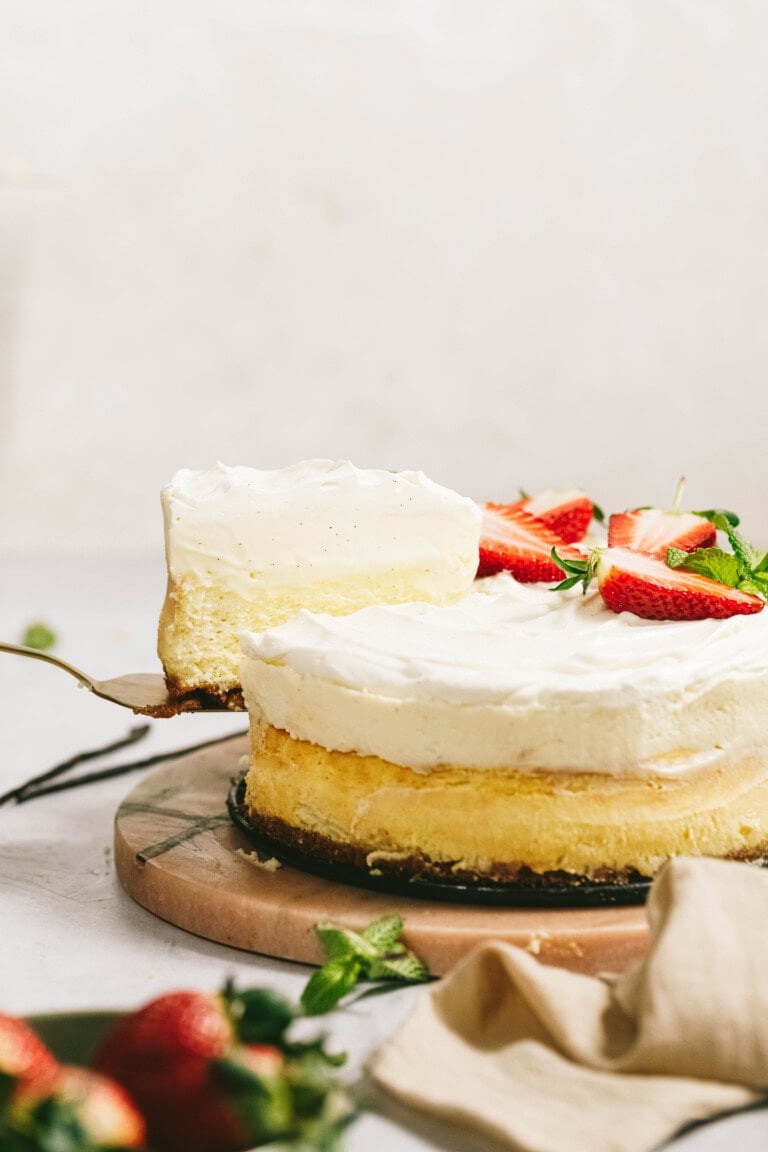 A slice of cream-covered cake is being lifted from a whole cake on a wooden board. The cake is topped with sliced strawberries and garnished with mint leaves.