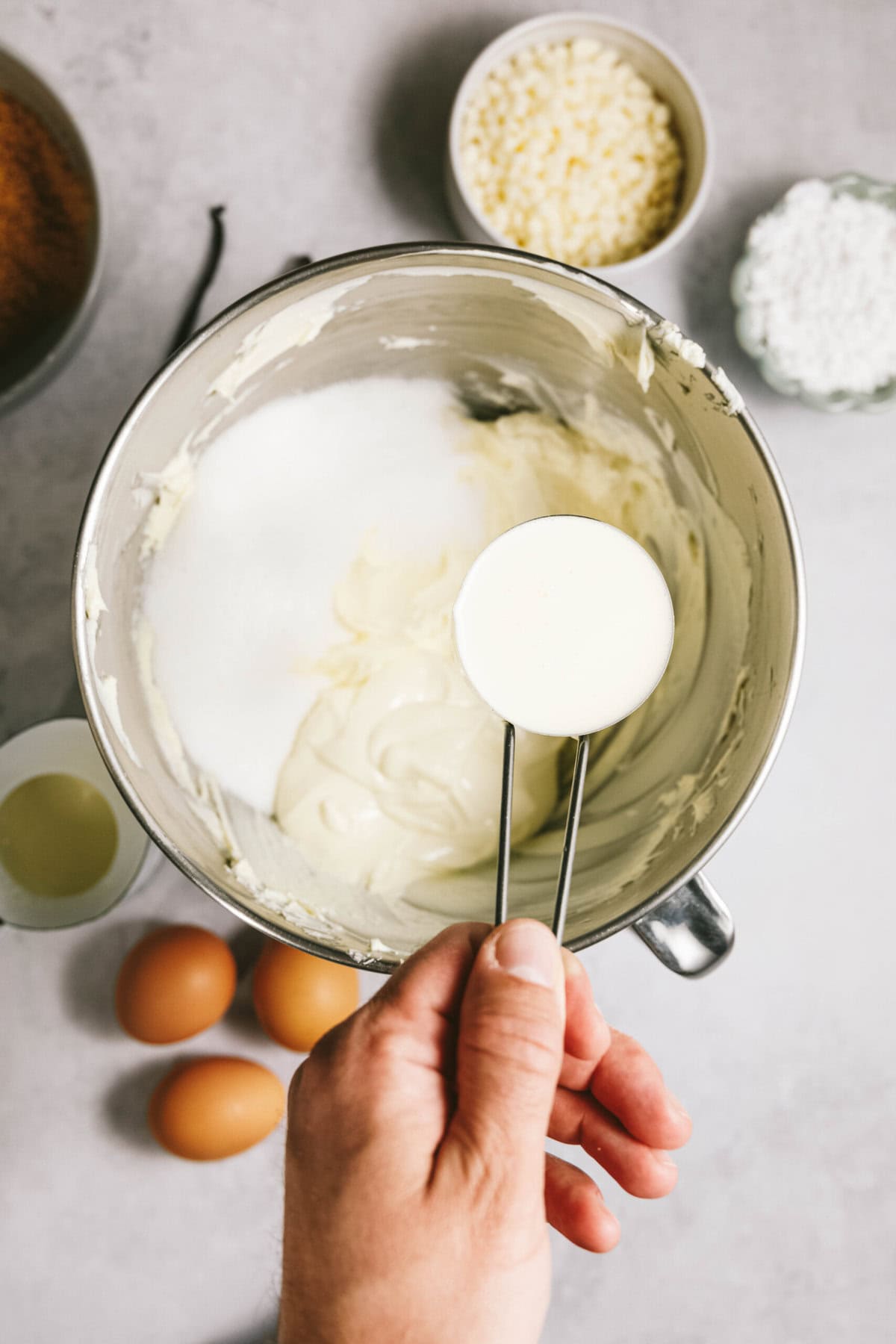 A hand holding a measuring spoon over a mixing bowl of batter. Nearby are three eggs, vanilla pods, a small bowl of white chocolate chips, and other ingredients.