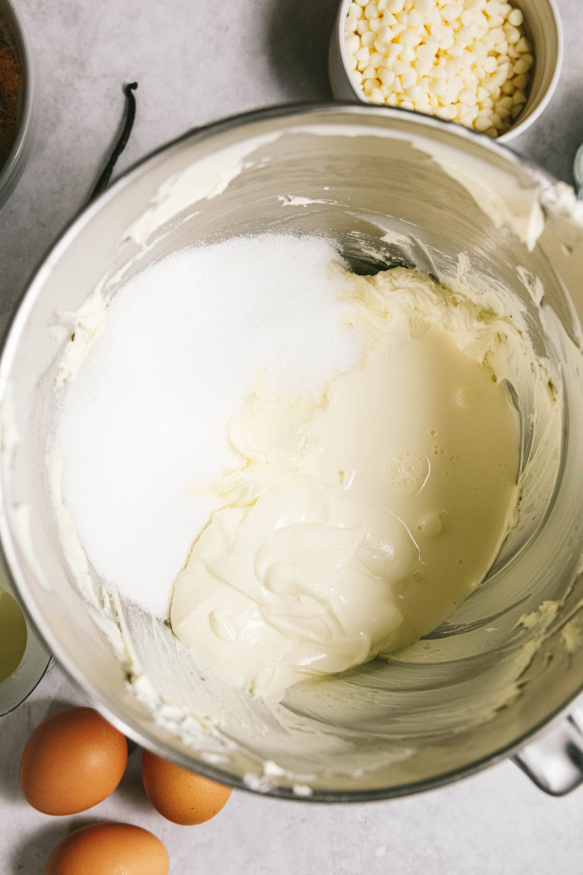 A mixing bowl containing a mixture of cream and sugar. Nearby are eggs, a bowl of white chocolate chips, and a few other ingredients.