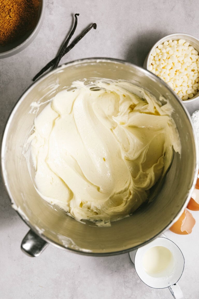 A mixing bowl filled with creamy white batter, surrounded by ingredients such as white chocolate chips, eggs, and vanilla beans on a gray countertop.