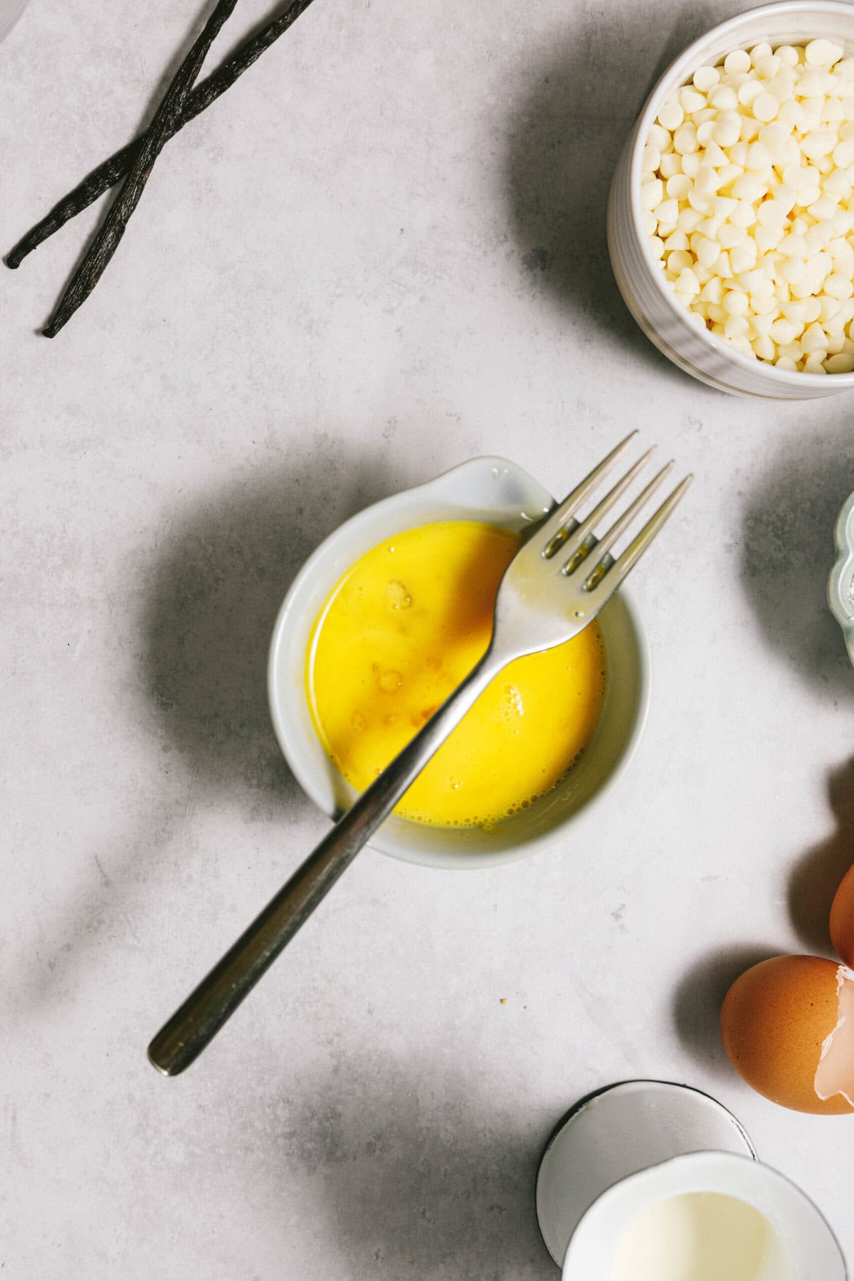 A bowl of beaten eggs with a fork, next to vanilla beans, white chocolate chips, two whole eggs, and a cup of cream on a light-colored surface.