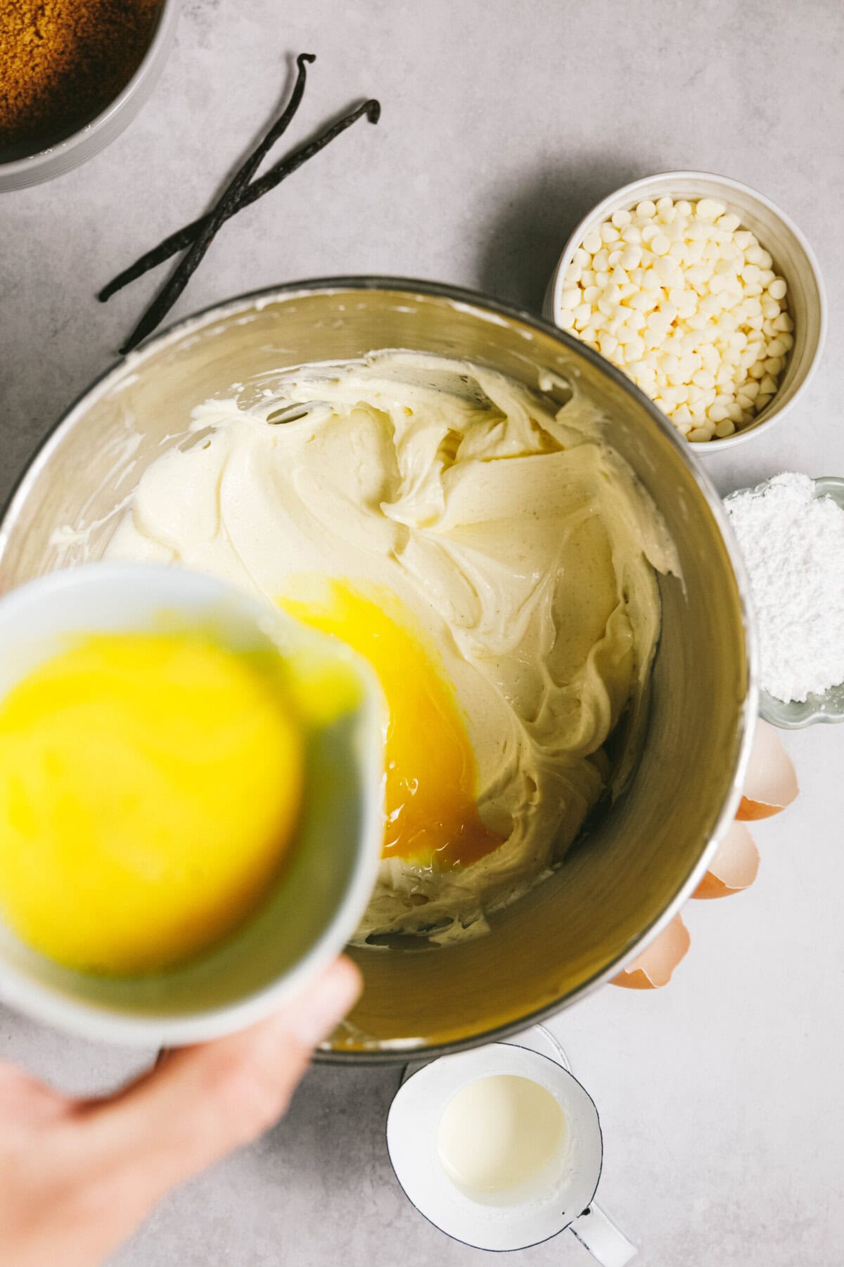 A bowl of creamy batter with an egg mixture being poured in, surrounded by ingredients like vanilla beans, white chocolate chips, powdered sugar, and a small cup of milk on a gray surface.