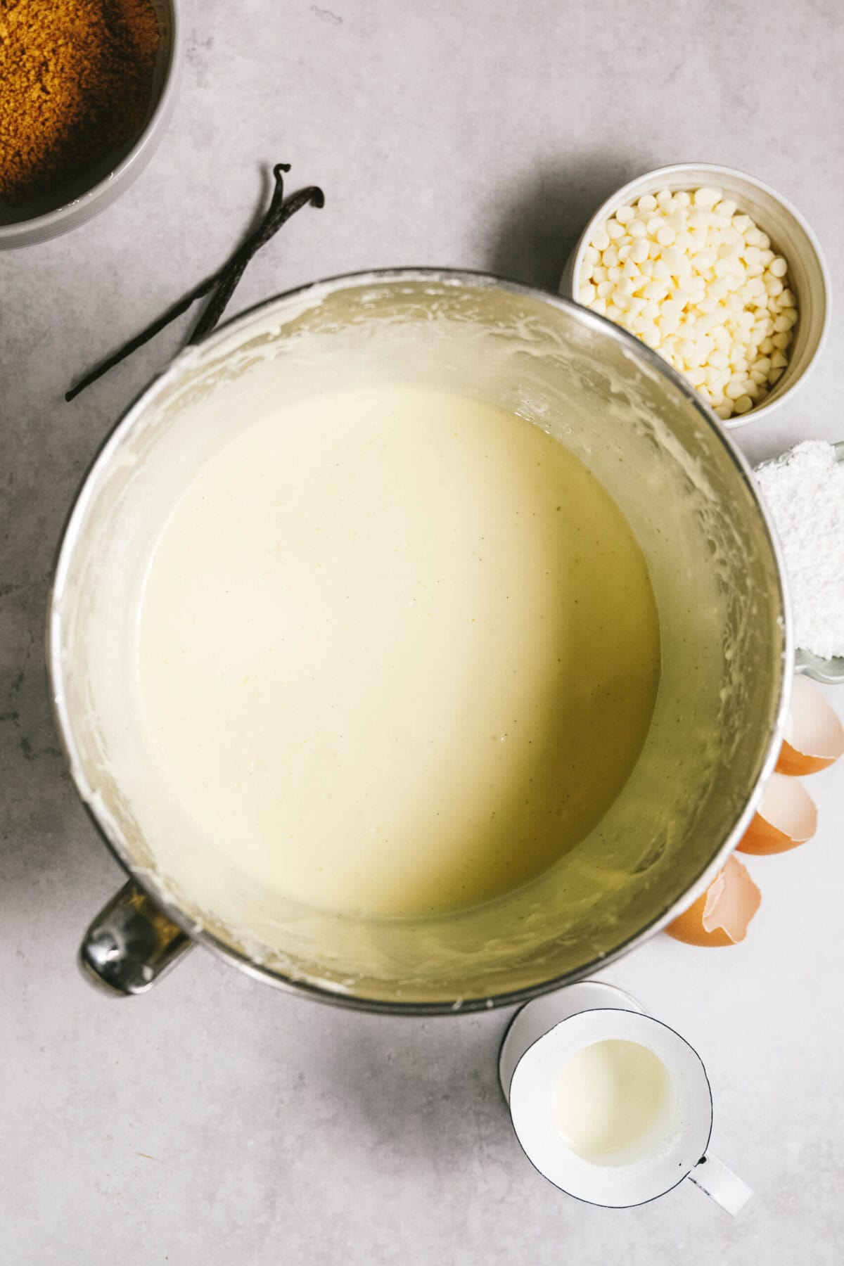 A mixing bowl filled with a creamy batter. Surrounding it are bowls of ingredients: crushed graham crackers, vanilla beans, white chocolate chips, powdered sugar, eggshells, and a small cup of cream.