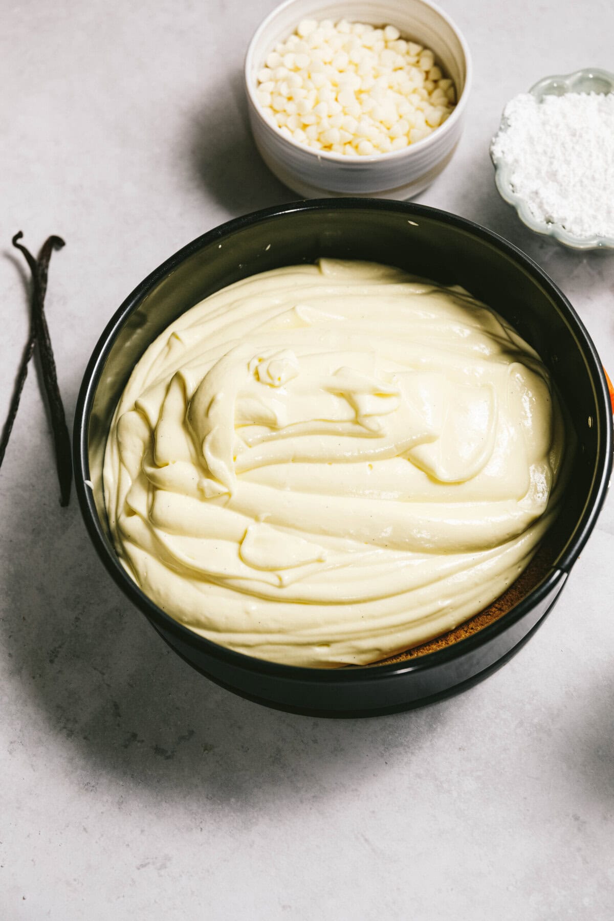 A bowl of creamy batter sits on a counter, surrounded by a vanilla pod, white chocolate chips, and a bowl of powdered sugar.