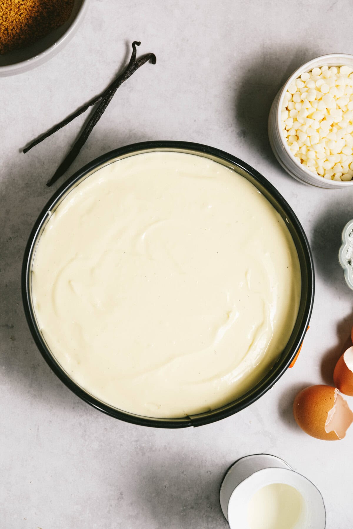 A round pan filled with smooth cheesecake batter. Nearby are vanilla beans, white chocolate chips, eggs, and a cup of cream.