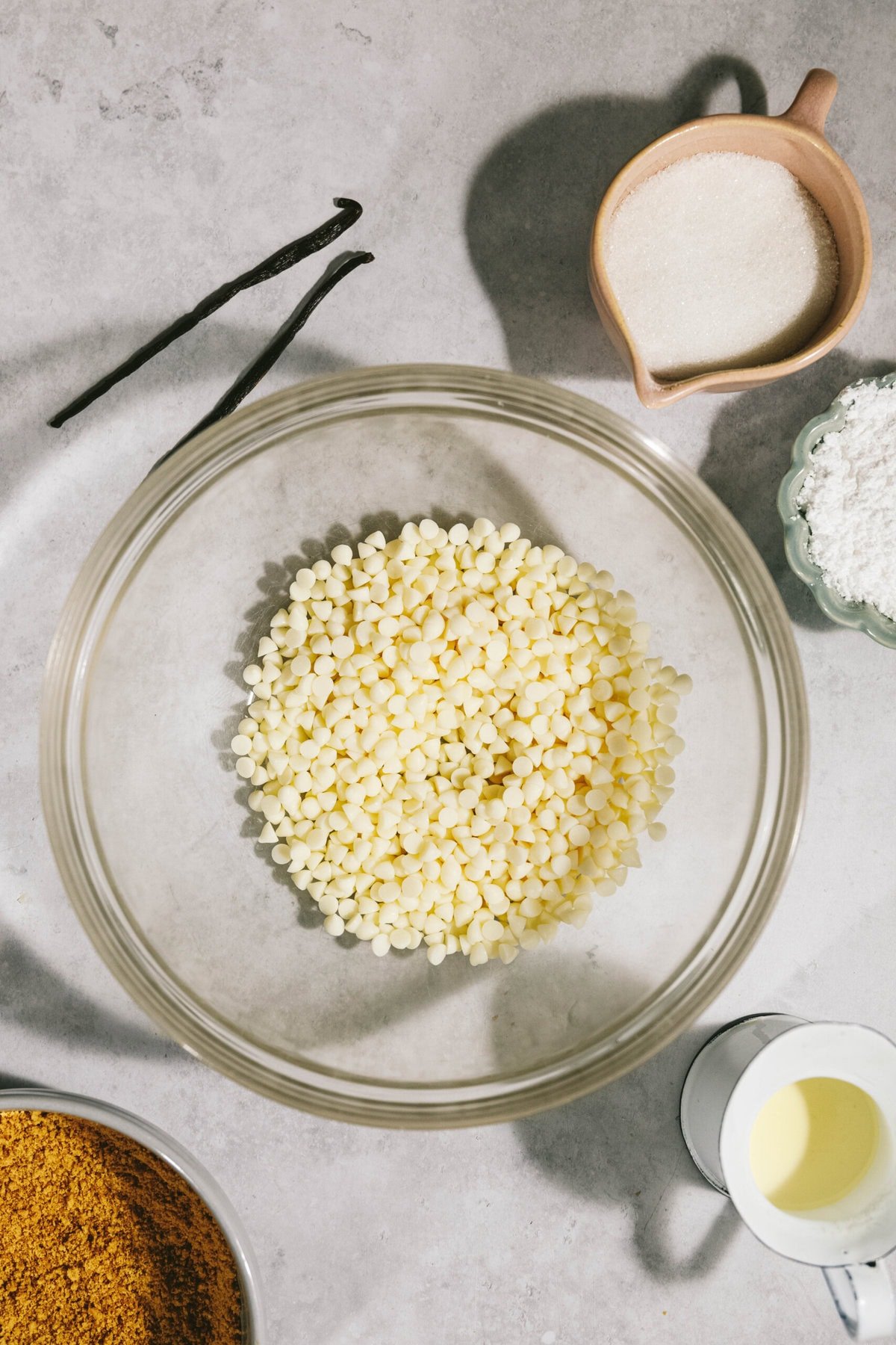 Top-down view of baking ingredients on a countertop, including a bowl of white chocolate chips, sugar, vanilla beans, and various powders, arranged in separate containers.