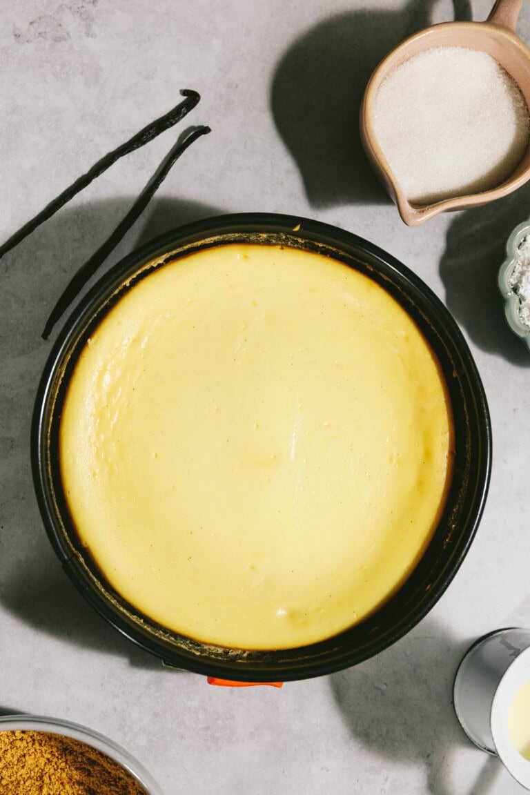 Overhead view of a round, freshly baked cheesecake in a springform pan, surrounded by vanilla beans, a bowl of sugar, and other baking ingredients on a gray surface.