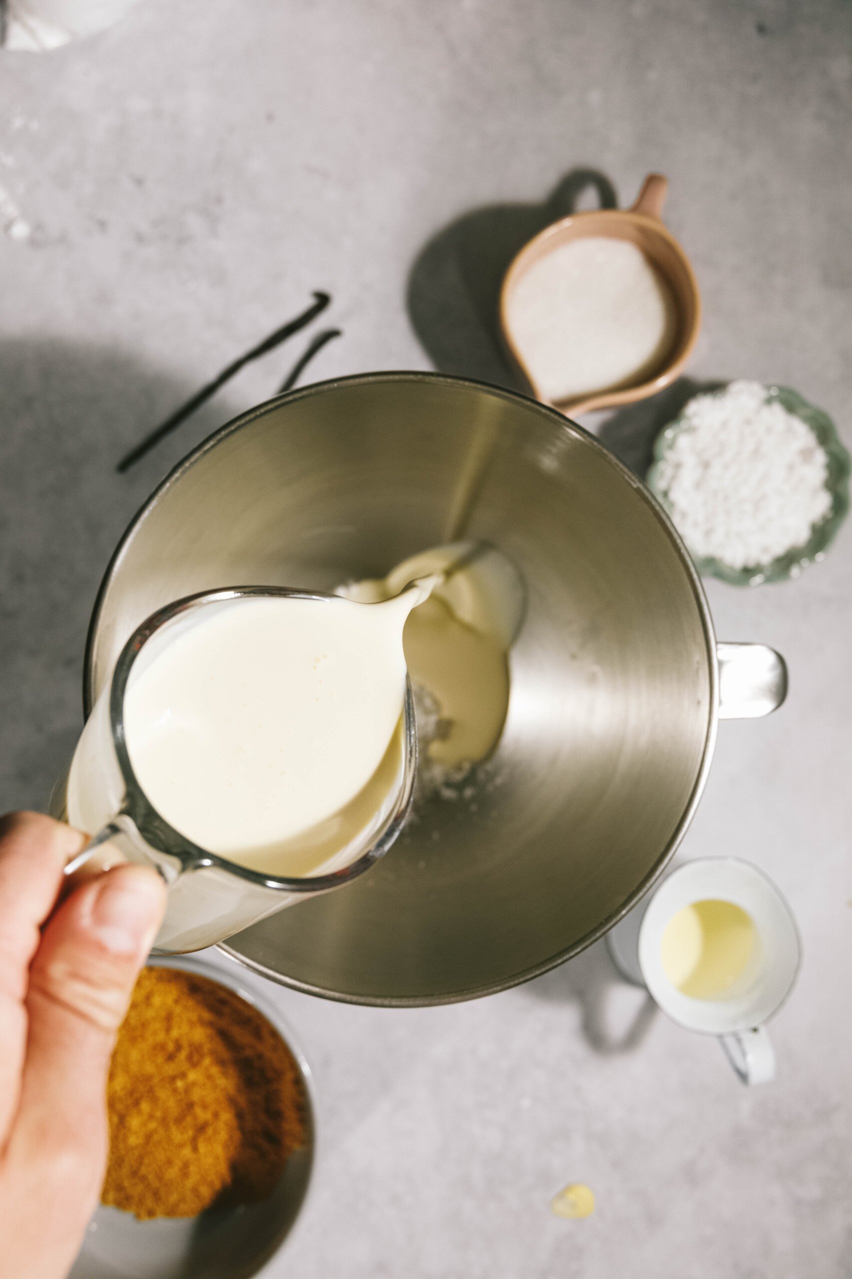 Person pouring cream into a mixing bowl, with various ingredients like brown sugar and flour arranged around it on a countertop.