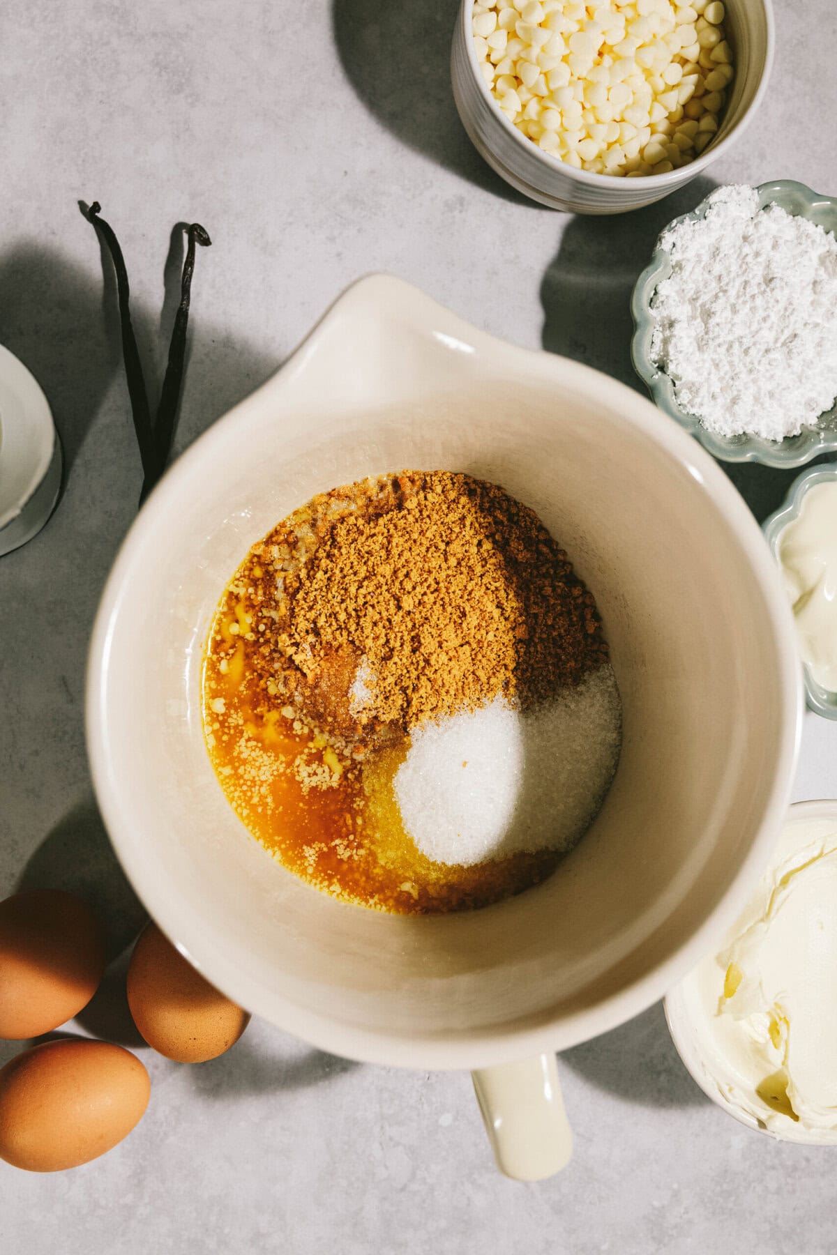 Mixing bowl with brown sugar, granulated sugar, and melted butter. Surrounding the bowl are eggs, vanilla beans, white chocolate chips, and powdered sugar.