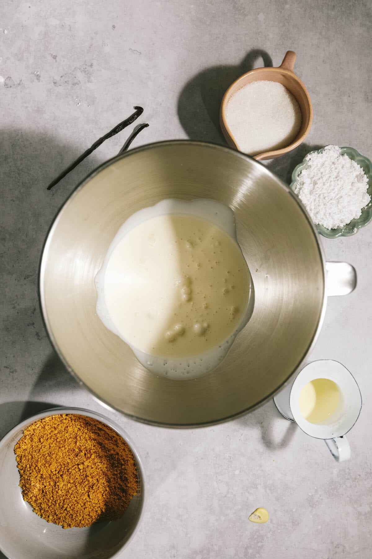 Ingredients laid out on a countertop for a recipe, including a bowl of milk, a cup of flour, a cup of sugar, a bowl of orange zest, a small pitcher of oil, a vanilla pod, and a clove of garlic.