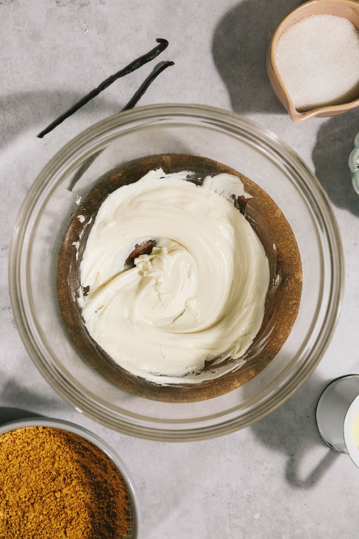 A bowl of creamy white frosting on a wooden surface, surrounded by a bowl of brown sugar, another bowl of granulated sugar, a pitcher of milk, and two vanilla beans.