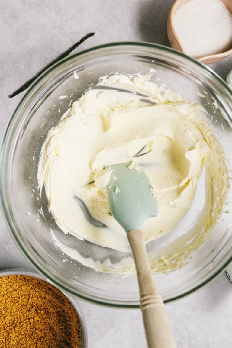 A glass bowl containing a creamy mixture being stirred with a green spatula. Surrounding the bowl are a container of brown crumbs, a dish of granular white substance, and a vanilla bean.