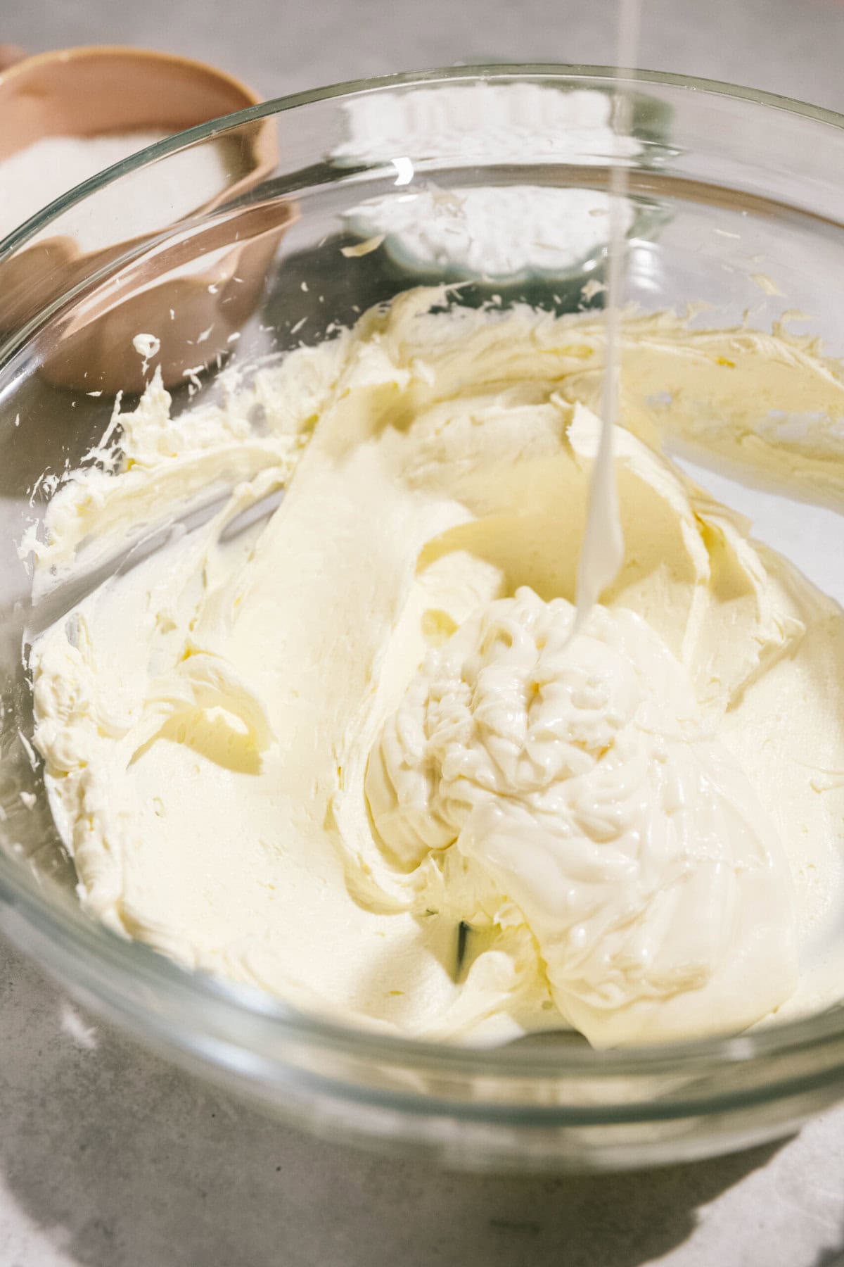 A glass bowl filled with creamy, light yellow batter or frosting. Liquid is being poured into the bowl from above. Two additional bowls with white and brown contents are in the background.