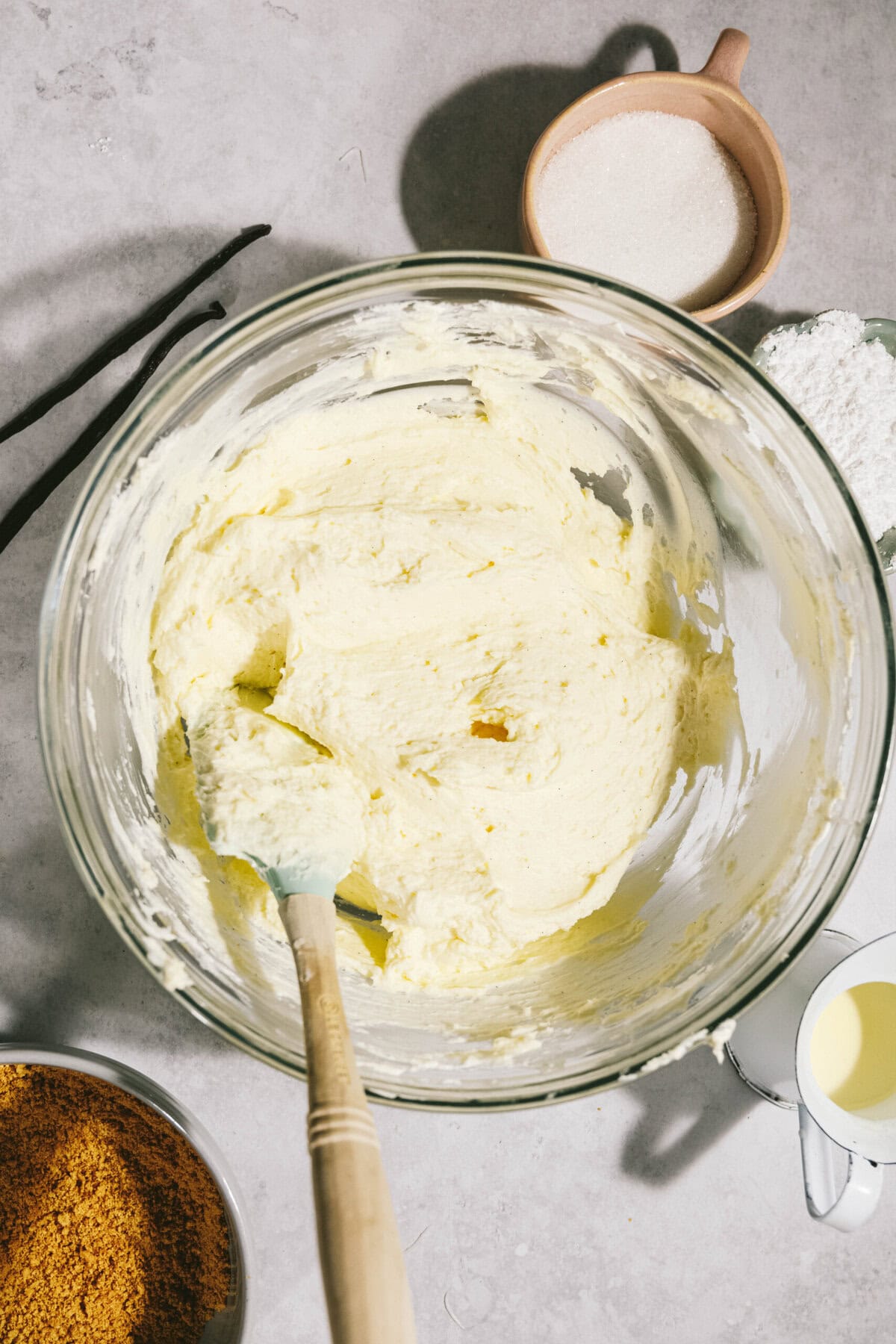 A glass bowl with creamy mixture and a spatula inside. Surrounding are a bowl of brown sugar, a small jug of liquid, a bowl of flour, a bowl of white granulated sugar, and two vanilla pods.