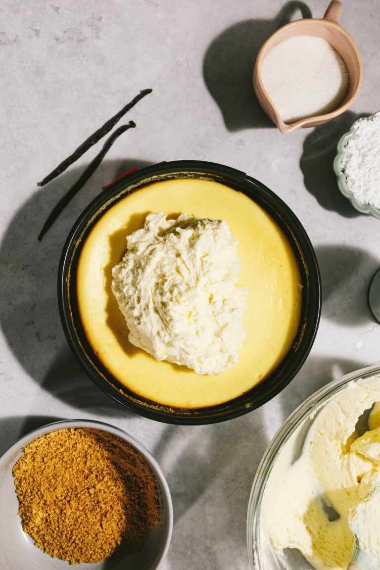 A lightly baked cheesecake topped with a large dollop of cream batter sits on a counter, surrounded by ingredients including vanilla beans, sugar, crushed crackers, and flour.