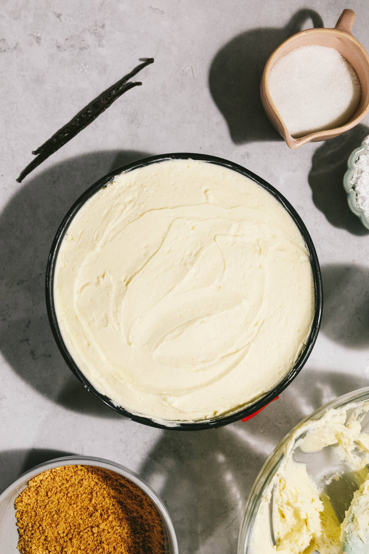 A bowl of smooth, pale yellow batter is surrounded by ingredients including two vanilla beans, a pitcher of sugar, and a bowl of crumbled crust mix on a gray countertop.