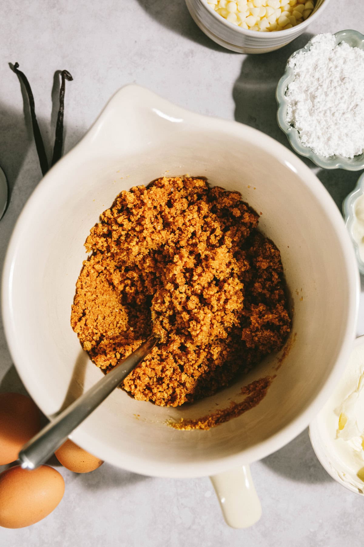 A mixing bowl with a crumbly, caramel-colored mixture and a spoon inside. Surrounding ingredients include eggs, vanilla beans, powdered sugar, white chocolate chips, and softened butter.