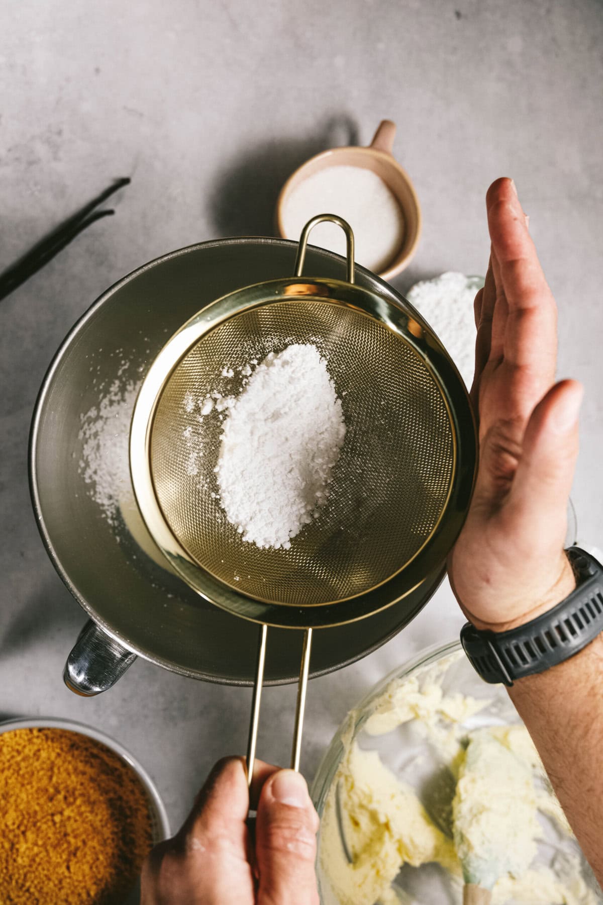 A person sifting powdered sugar through a metal sieve over a mixing bowl on a gray countertop, with various ingredients around.