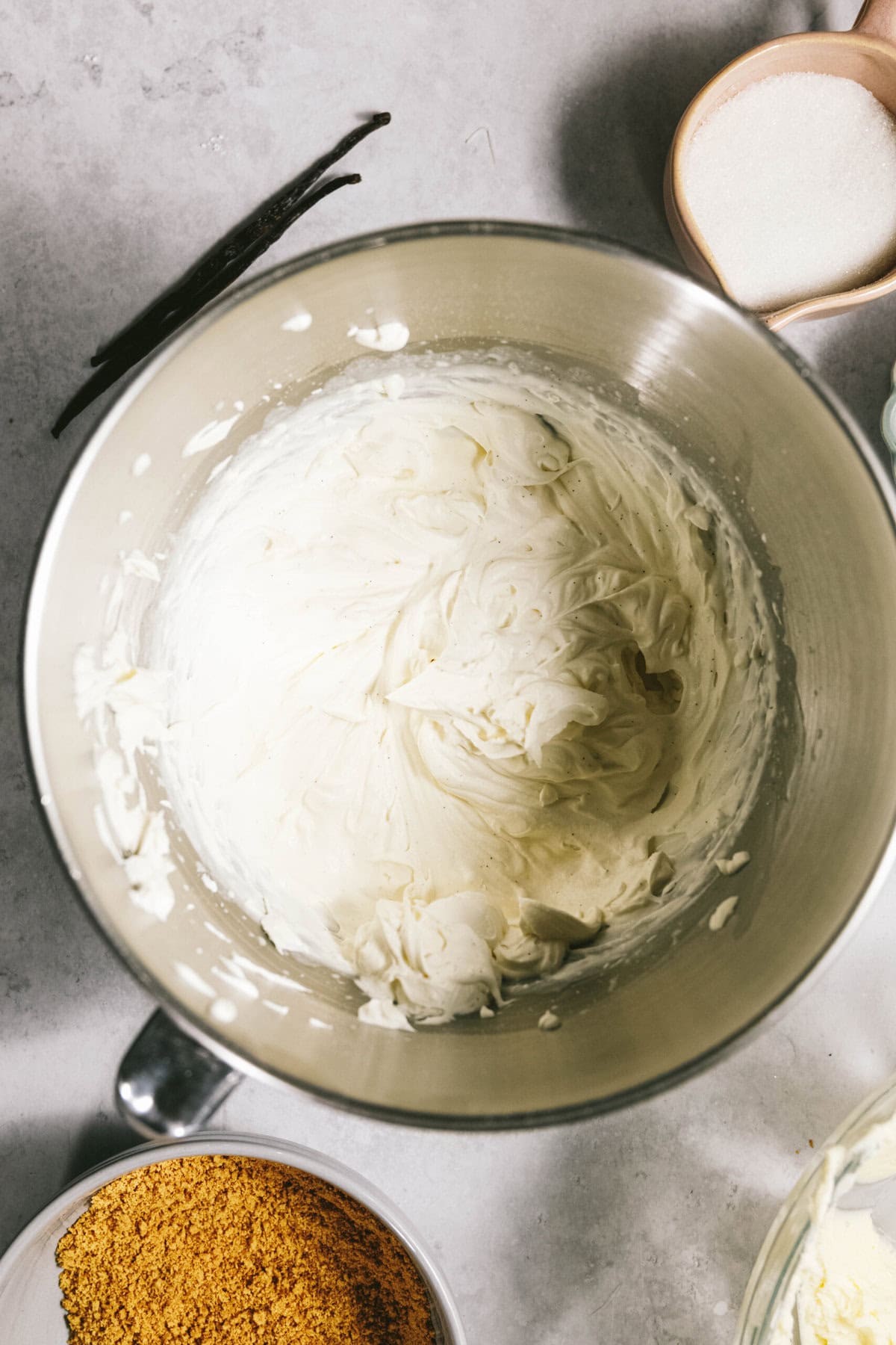 A mixing bowl containing whipped cream cheese mixture, surrounded by a small bowl of sugar, a small bowl of crushed graham crackers, and two vanilla beans on a light gray surface.