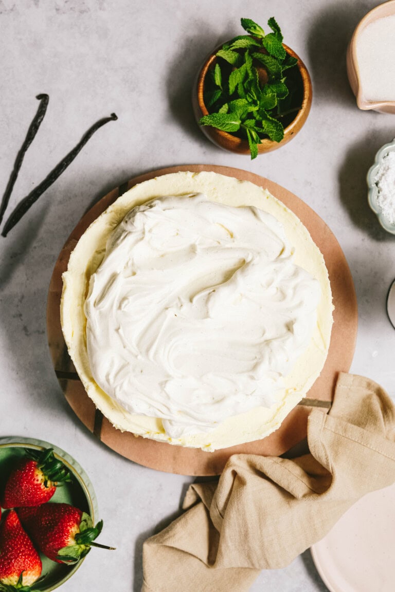 A round cake topped with frosting sits on a wooden platter, surrounded by fresh strawberries, a bowl of mint leaves, vanilla beans, and a beige cloth on a light-colored surface.