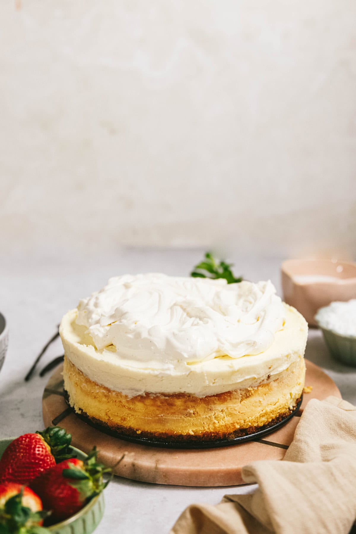 A cheesecake with a layer of whipped cream on a wooden board, with strawberries and a small bowl of powdered sugar in the foreground.