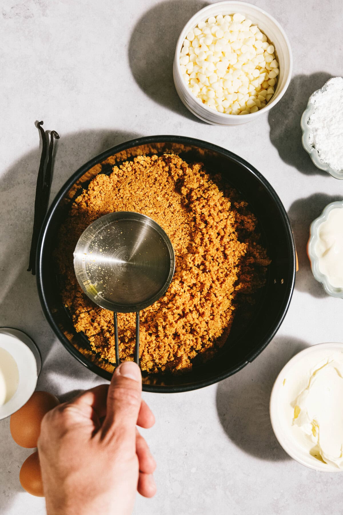 A hand holding a measuring cup over a bowl of crumb mixture, surrounded by ingredients including white chocolate chips, eggs, vanilla beans, butter, and powdered sugar on a counter.