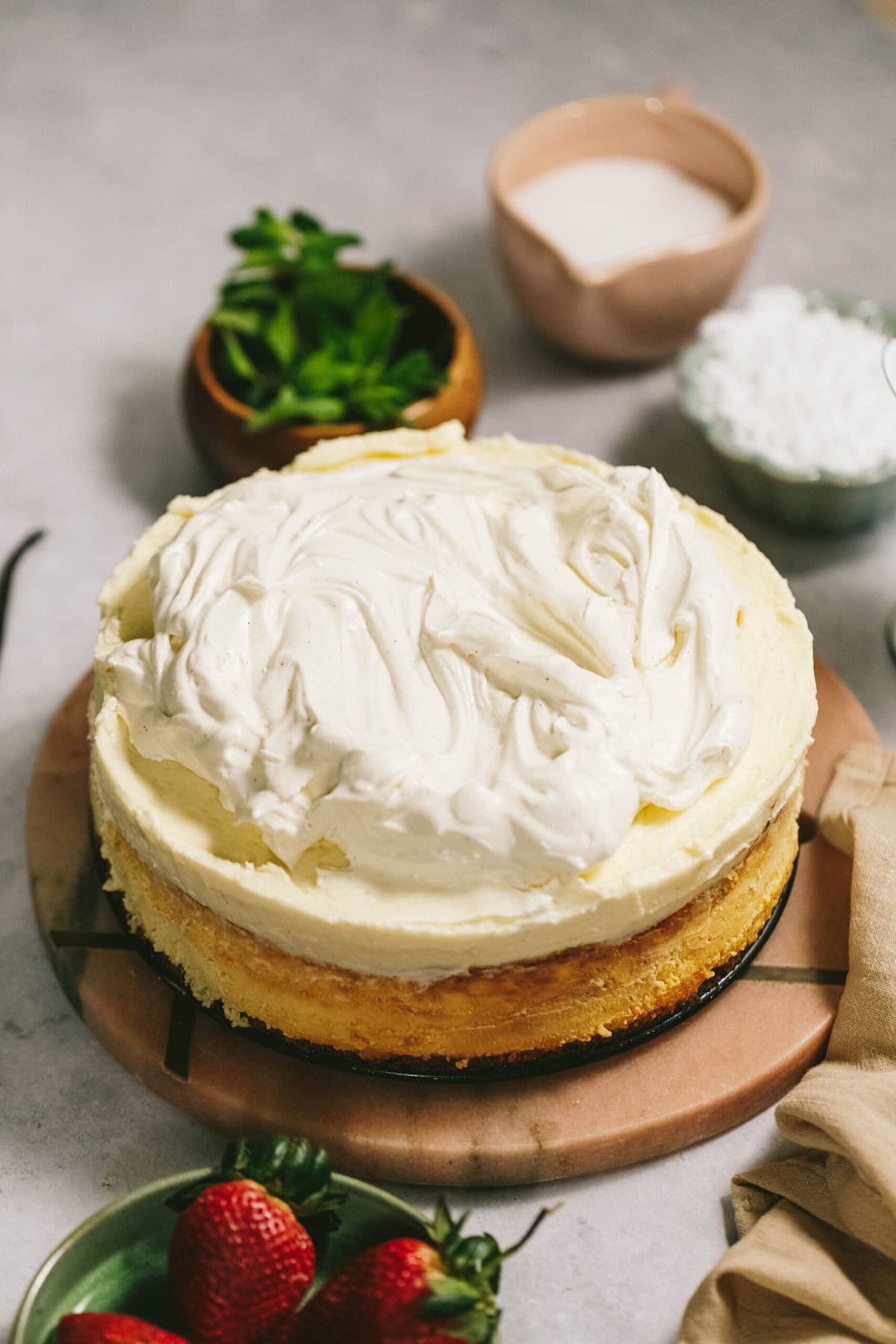 A frosted cake on a round board with mint leaves, a small bowl of icing, a bowl of powdered sugar, and a plate of strawberries arranged around it.