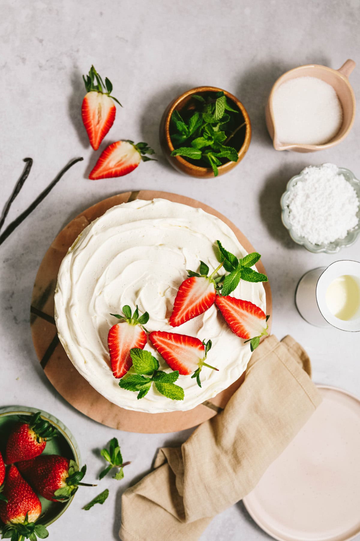 A frosted cake topped with strawberries and mint leaves is displayed on a wooden board with scattered strawberries, mint, sugar, and a beige napkin.