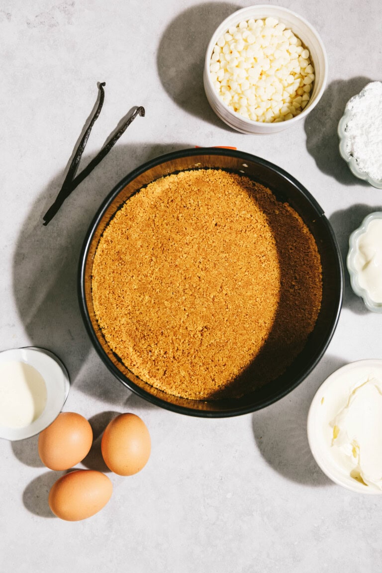 Ingredients for a baking recipe laid out on a surface, including a pie crust in a pan, three eggs, vanilla beans, white chocolate chips, powdered sugar, cream, and cream cheese.