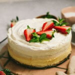 A round cheesecake topped with white frosting, fresh strawberries, and mint leaves, placed on a pinkish-brown surface.