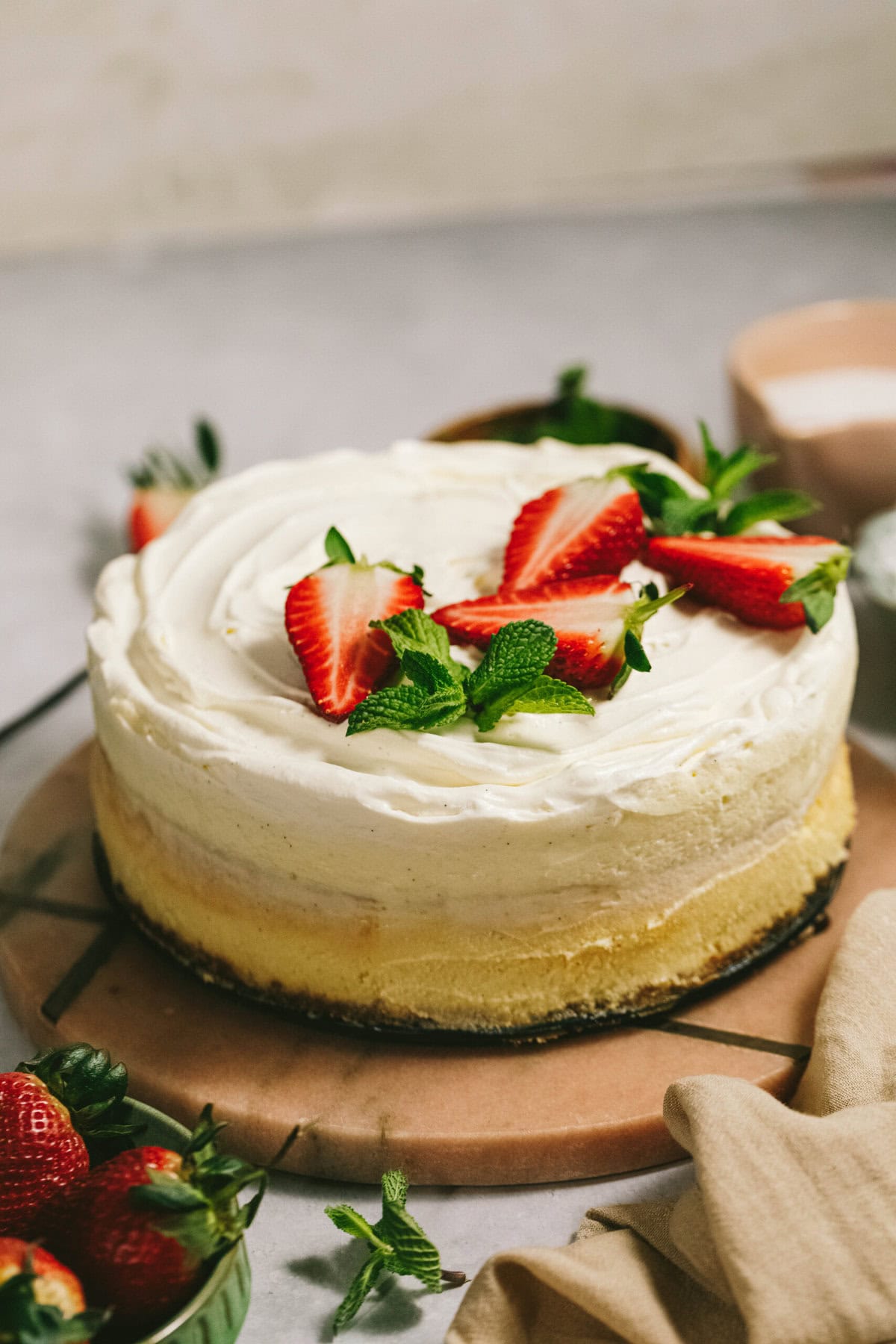 A round cheesecake topped with white frosting, fresh strawberries, and mint leaves, placed on a pinkish-brown surface.