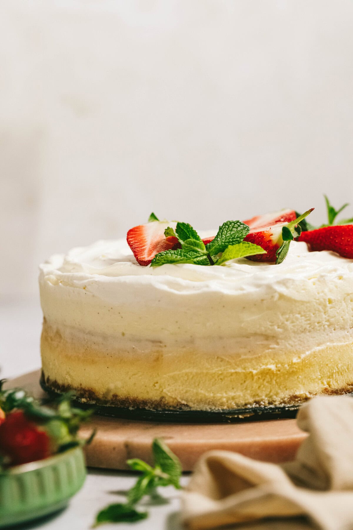 A cheesecake topped with fresh strawberry slices and mint leaves on a light-colored surface with a green bowl and beige cloth nearby.