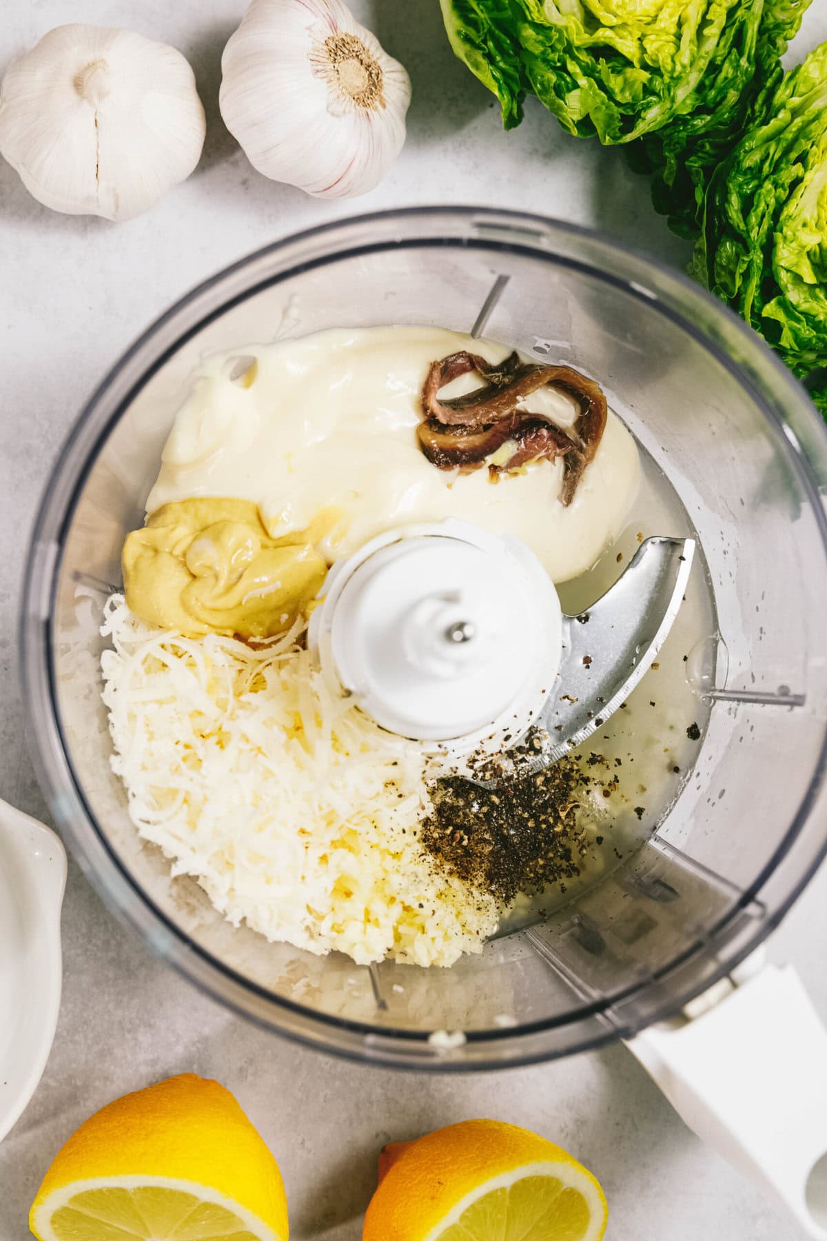 Food processor containing ingredients including mayonnaise, garlic, mustard, Parmesan cheese, anchovies, lemon juice, and black pepper, surrounded by garlic bulbs, lemon halves, and lettuce.