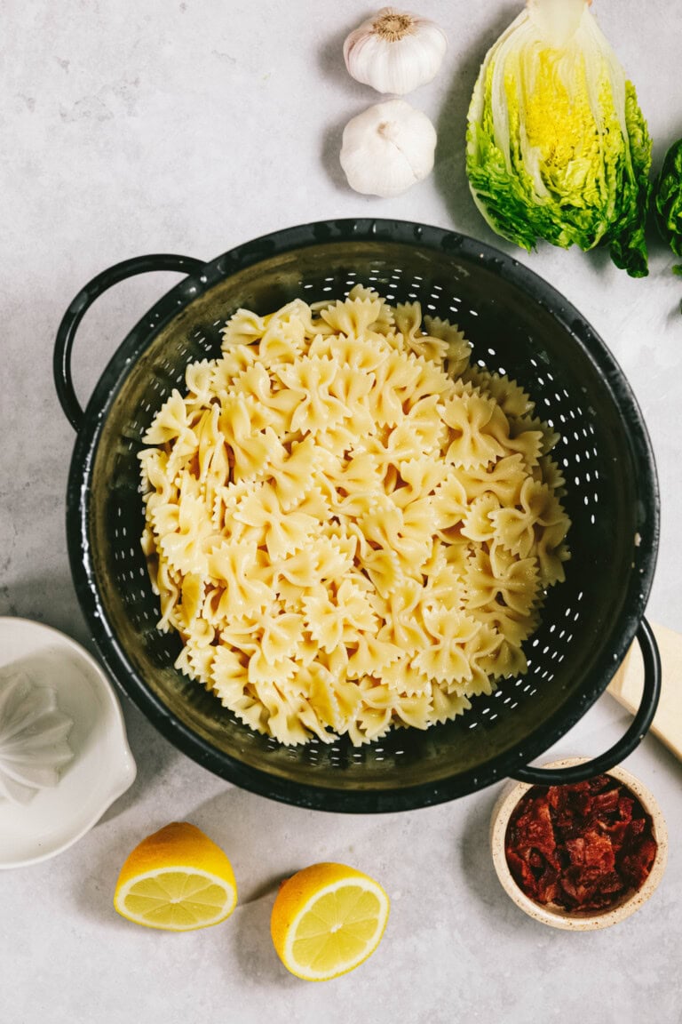 A black colander filled with cooked farfalle pasta, surrounded by lemon halves, sun-dried tomatoes, garlic cloves, and romaine lettuce on a light-colored surface.