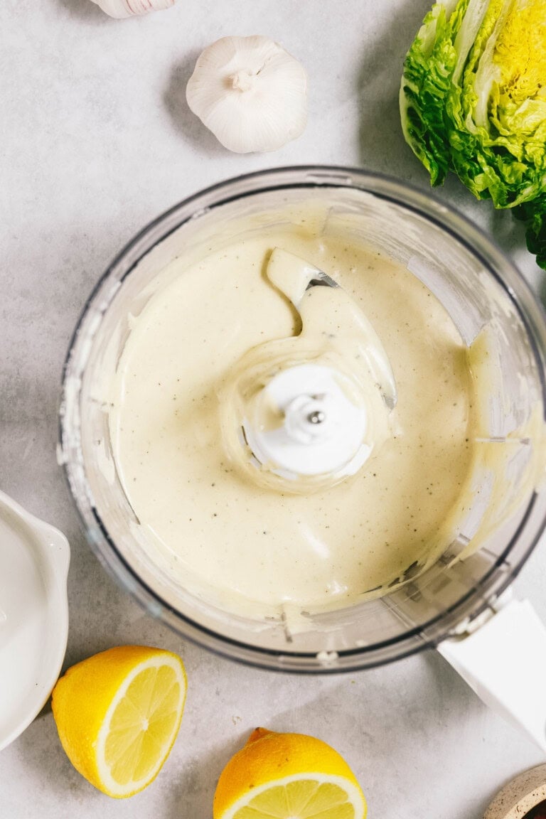 A top-down view of a food processor containing creamy dressing. Surrounding it are garlic bulbs, a halved lemon, a whole lemon, and a small lettuce head on a light gray surface.