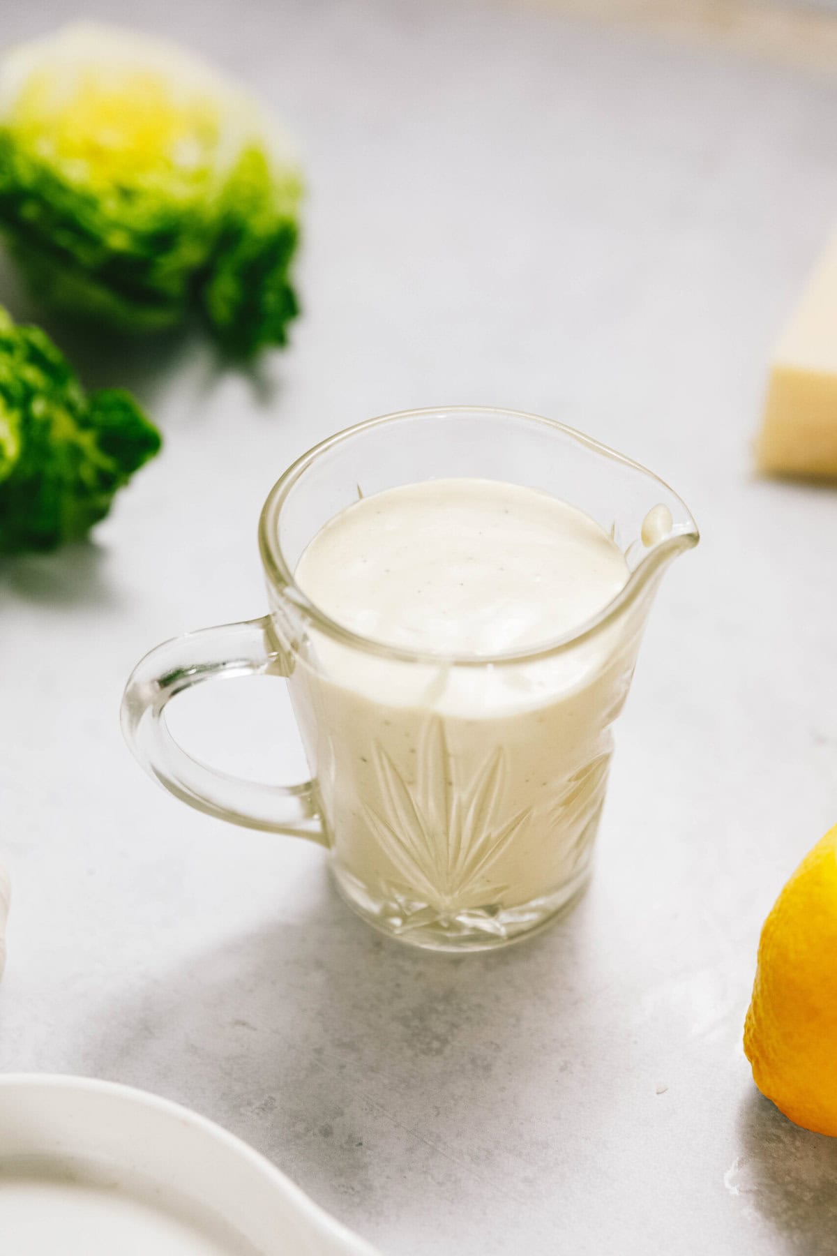 A glass pitcher filled with white creamy dressing sits on a light-colored surface. Fresh lettuce and a lemon are nearby.