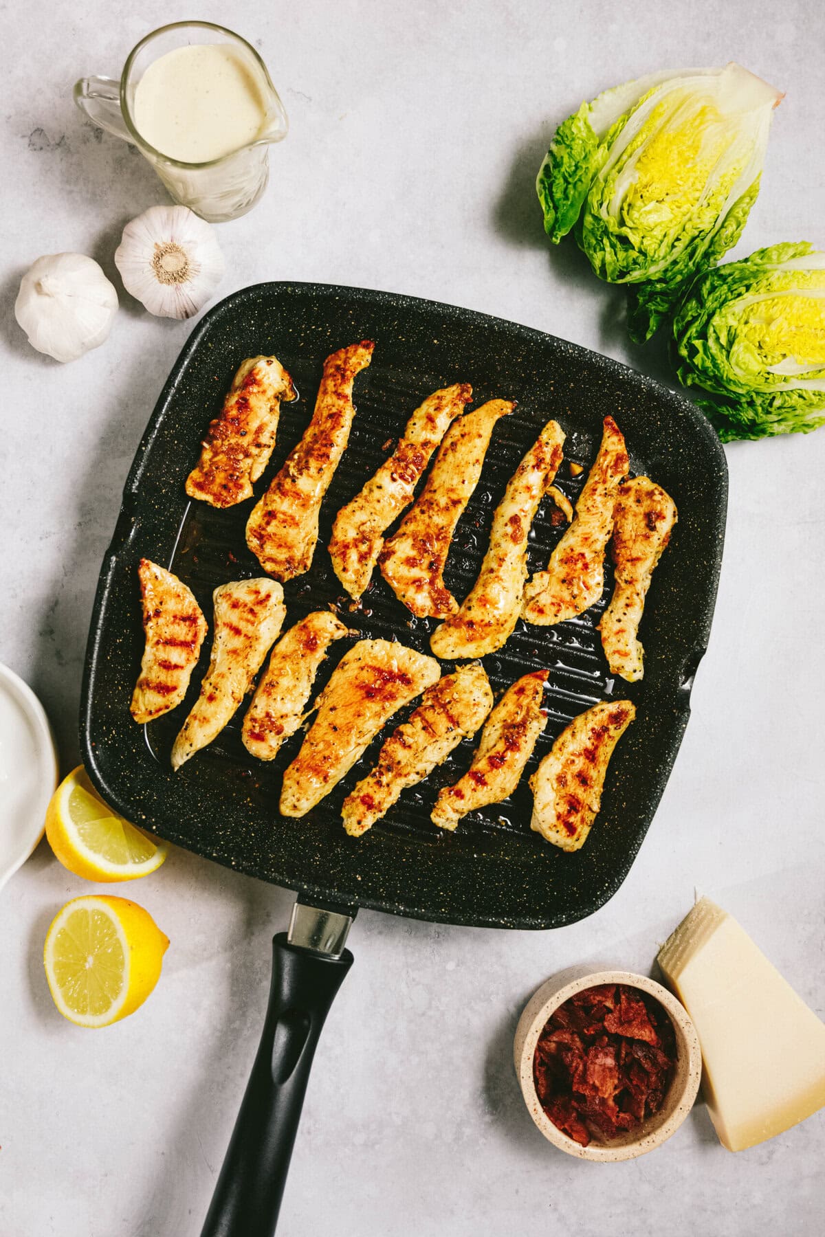 Grilled chicken strips in a black pan surrounded by garlic, romaine lettuce halves, Caesar dressing, lemon slices, sundried tomatoes, and cheese on a light countertop.