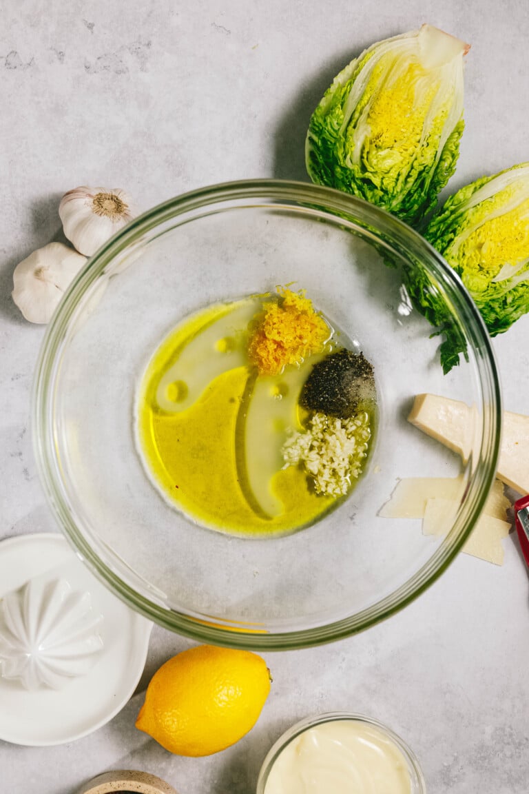 A glass bowl with olive oil, lemon zest, black pepper, and minced garlic. Surrounding the bowl are romaine lettuce, garlic, a lemon, a lemon juicer, and Parmesan cheese.