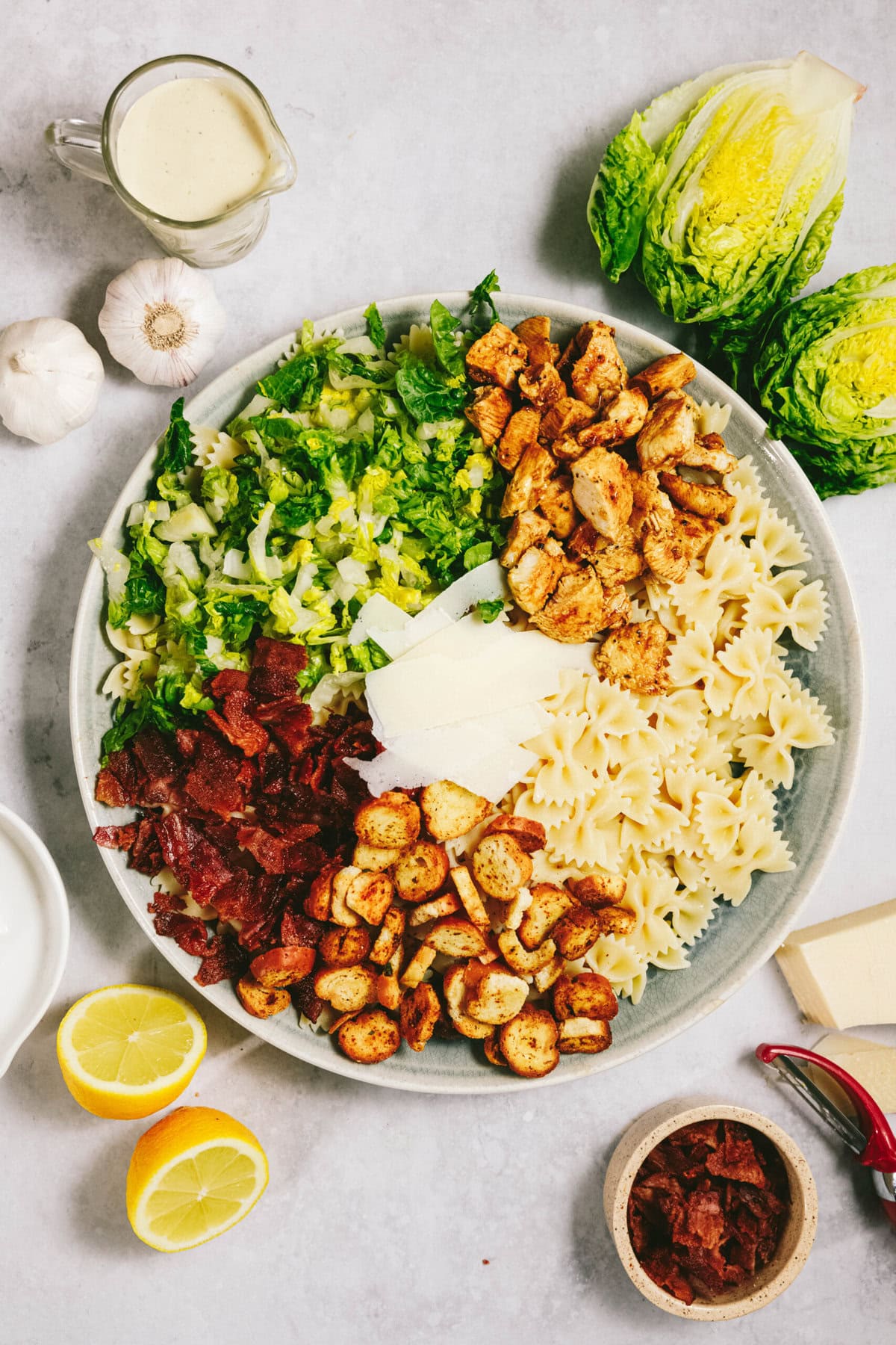 A bowl filled with chopped lettuce, grilled chicken, bow-tie pasta, bacon bits, croutons, and Parmesan cheese, with garlic, lemon halves, a sauce jug, and lettuce heads placed around it.