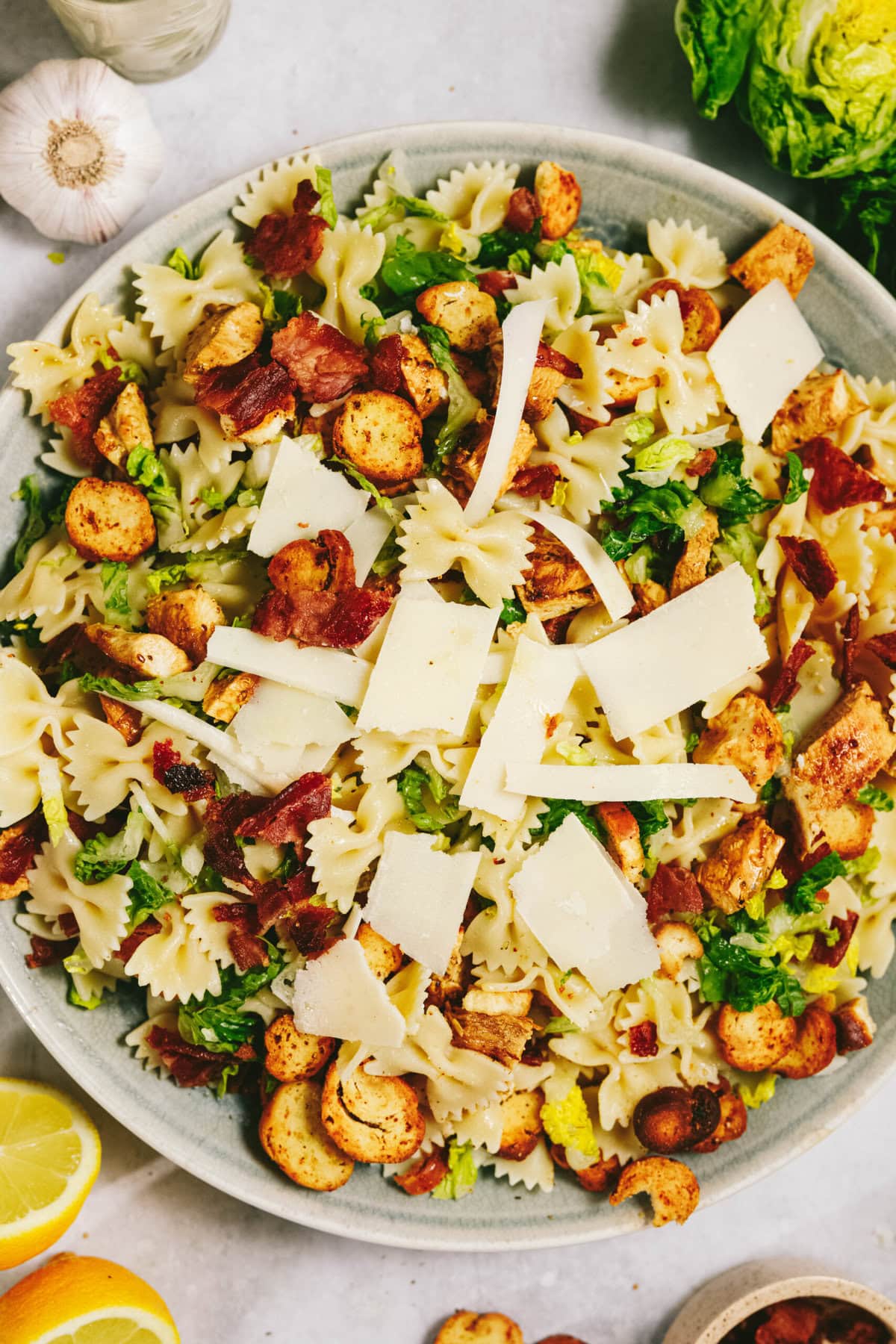 A bowl of bowtie pasta salad topped with croutons, bacon pieces, fresh greens, and large shavings of Parmesan cheese. Garlic and lemon wedges are visible in the background.