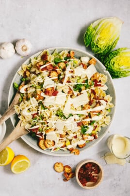 A bowl of pasta salad topped with croutons, sliced vegetables, and grated cheese, with lemon halves, garlic, lettuce, and a small bowl of dressing on the side.