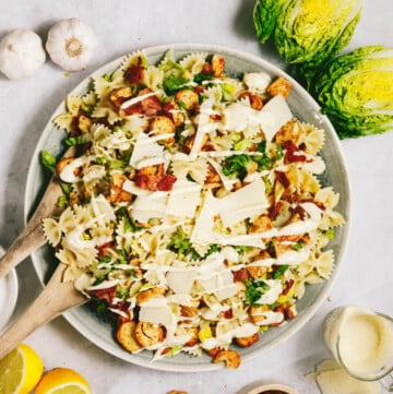 A bowl of pasta salad topped with croutons, sliced vegetables, and grated cheese, with lemon halves, garlic, lettuce, and a small bowl of dressing on the side.