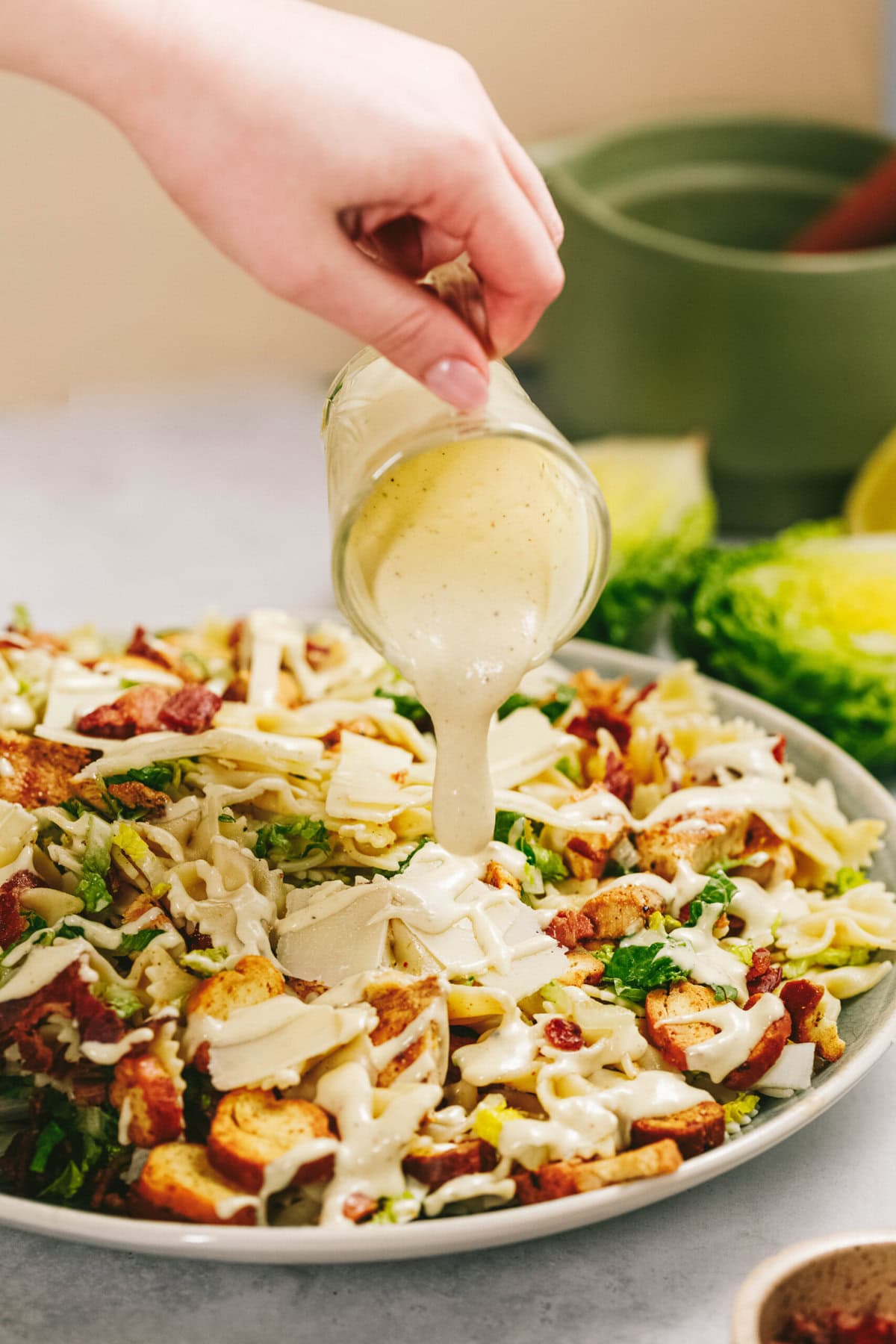 A hand pours creamy dressing from a small glass jar over a bowl of salad topped with chicken, cheese, and bacon.